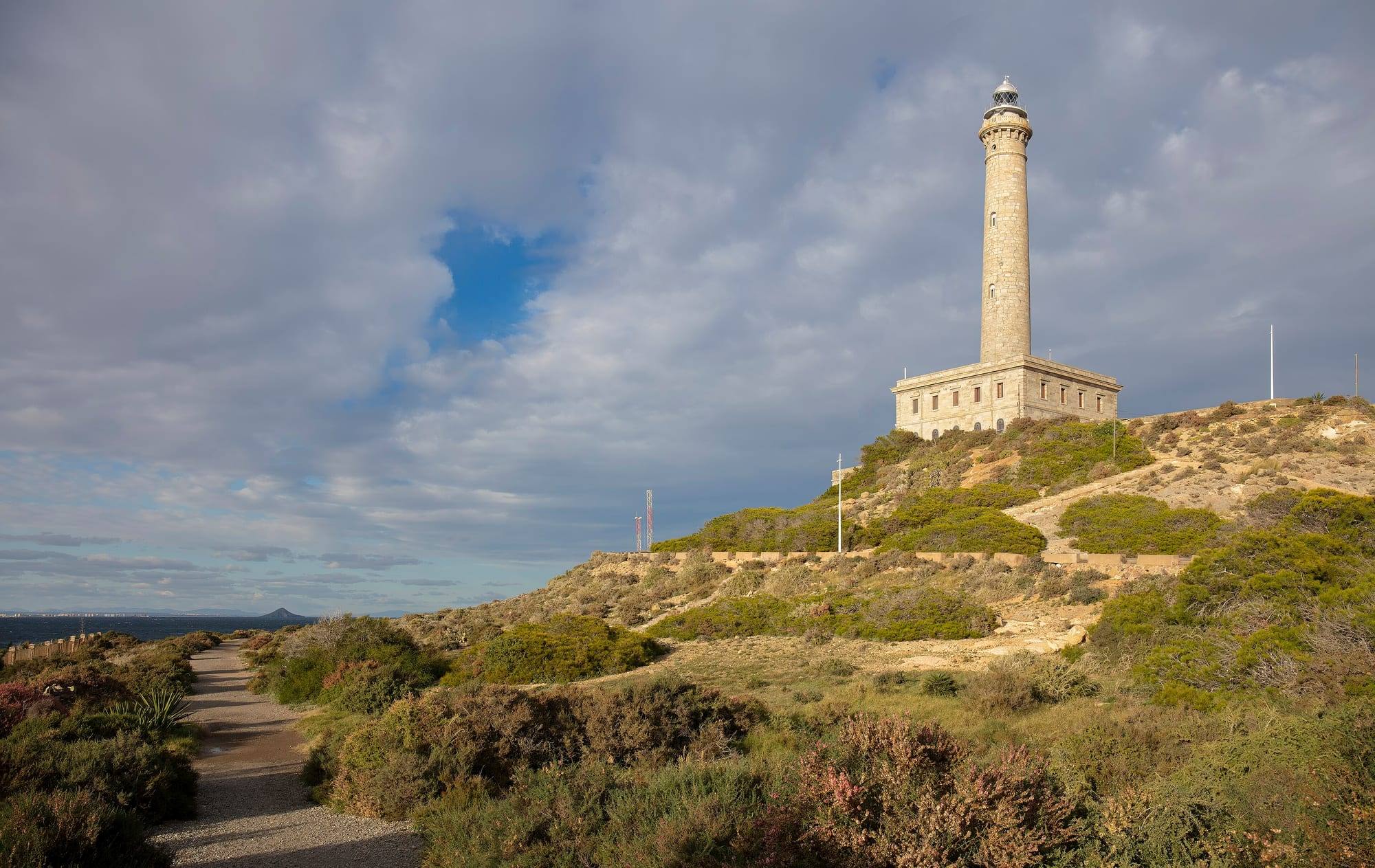 The Cabo de Palos lighthouse, a tall stone tower rising from a square two-storey keeper's house on a hill of scrub and pink heather lit by low autumn sun, a gravel path running along the cape on the left towards the dark Mediterranean, a distant island and the towers of La Manga on the horizon, under a broad grey-and-white cloudy sky with one patch of blue